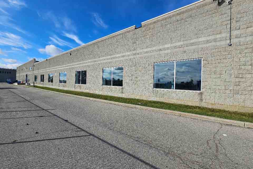 Side angled view of a long low-rise commercial concrete block facility showing a line of large glass windows reflecting the sky next to a paved driving lane.