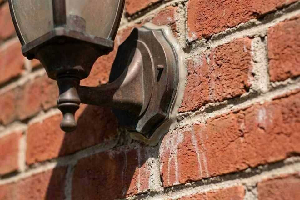 Close-up architectural view of a dark bronze carriage lamp baseplate flawlessly weatherproofed against a residential brick facade with a grey silicone bead.