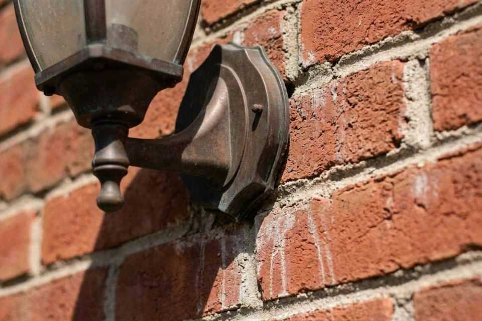 Close-up 4:3 field photograph of an outdoor bronze carriage lamp on a red brick wall with all old white caulking completely extracted, exposing a clean, raw joint channel.