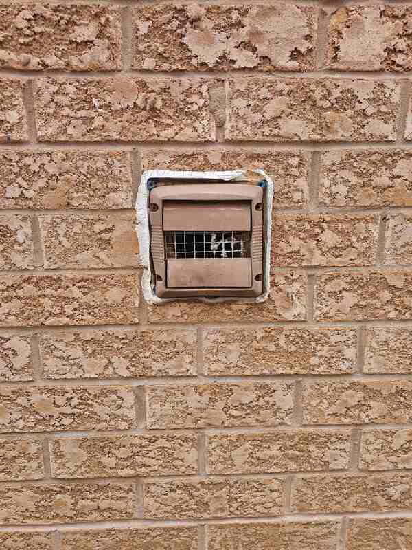 Close-up view of a brown plastic dryer vent exhaust cap on a tan brick wall showing thick, cracked, peeling white caulking separating from the housing flange.