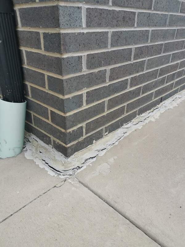 Close-up of a cracked and split horizontal expansion joint sealant line between a brick house foundation wall and a concrete sidewalk.