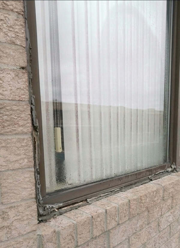 Close-up of a failed window seal with cracked caulking and trapped moisture condensation between double pane glass on a brick house in Burlington.