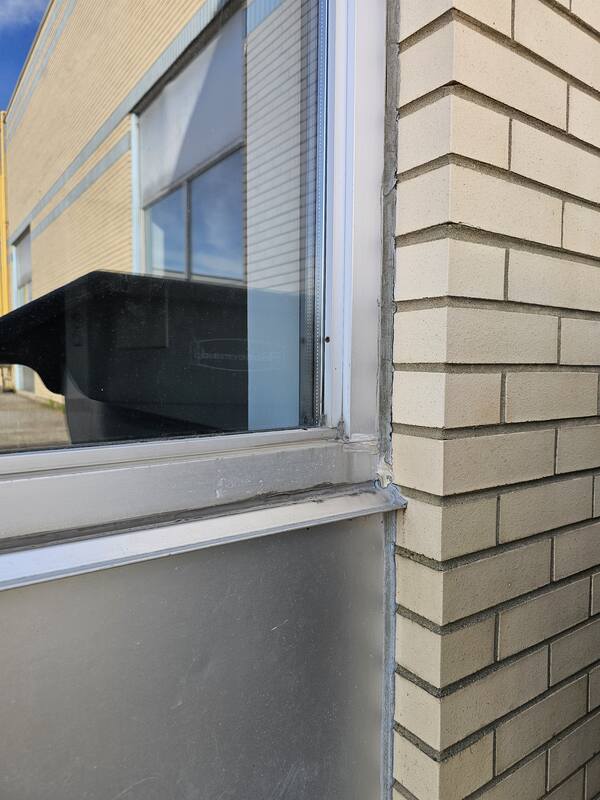 Close-up of dried, cracked, and shrinking old grey caulking sealant pulling away from the joint line between a commercial aluminum window frame and a brick wall facade.