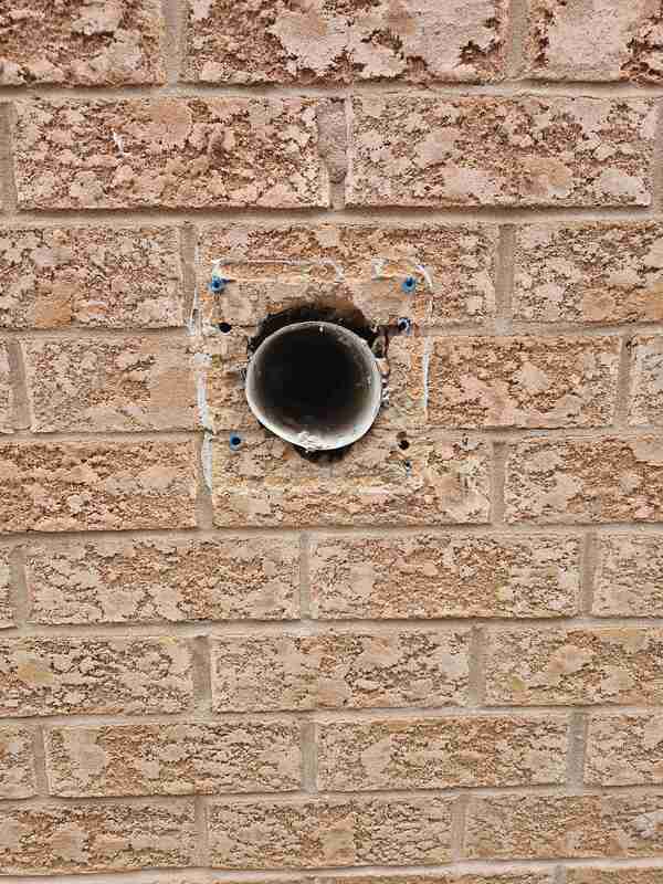 Close-up view of a raw circular exhaust pipe penetration on a tan brick wall with the plastic vent hood removed for substrate preparation.