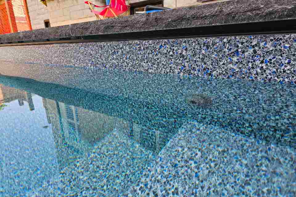 Wide angled view of a crystal clear inground swimming pool showing a long, uniform line of black silicone sealant perfectly applied under a dark stone coping ledge.