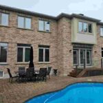 Rear exterior view of a two-story brick home in Burlington featuring a swimming pool, patio, and newly caulked tan windows by Proper Caulking.