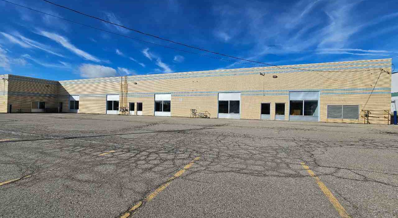 Wide exterior view of a low-rise commercial brick plaza and retail business center with glass windows, storefront doors, and a large paved parking lot under a blue sky.
