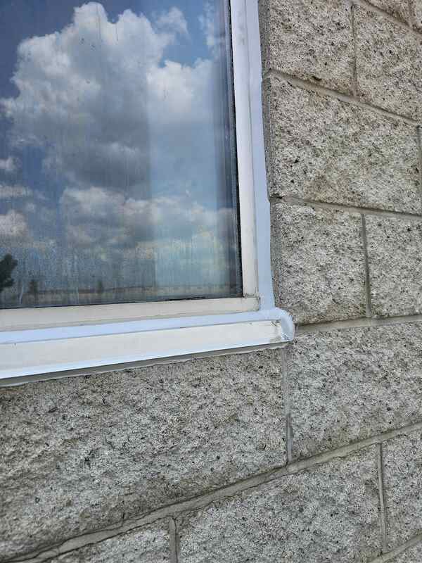 Close-up of a perfectly smooth, newly applied grey silicone caulking joint sealing the interface between a metal window frame and a rough split-face concrete block wall.
