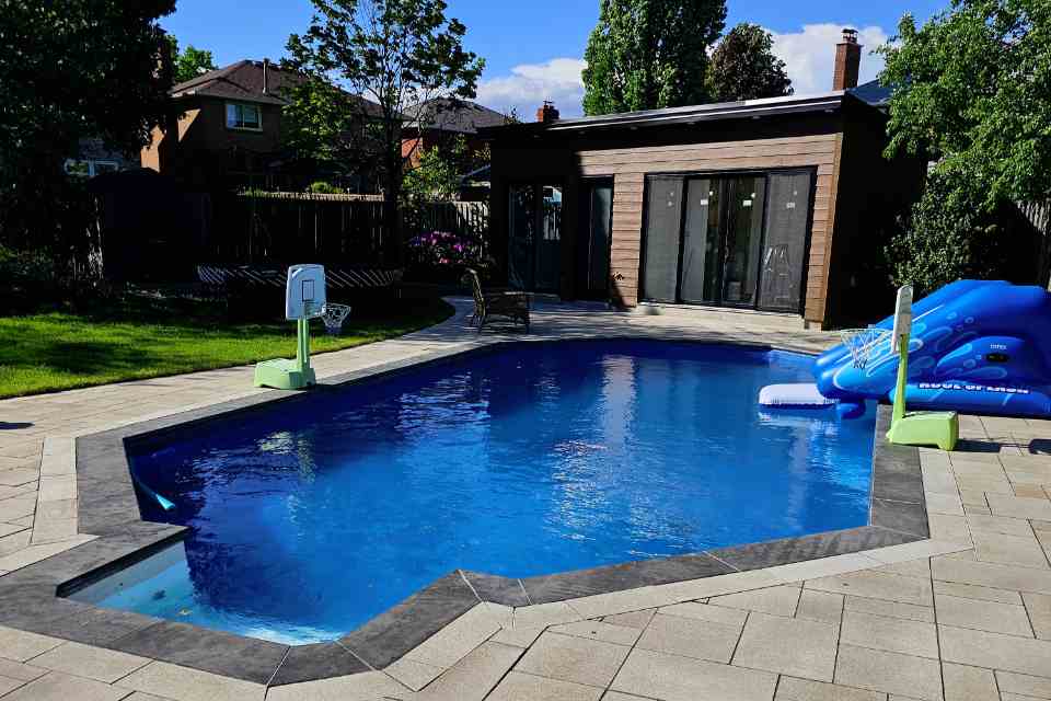 Wide view of a completed luxury inground swimming pool with finished dark stone coping expansion joints and an interlocking stone patio layout.