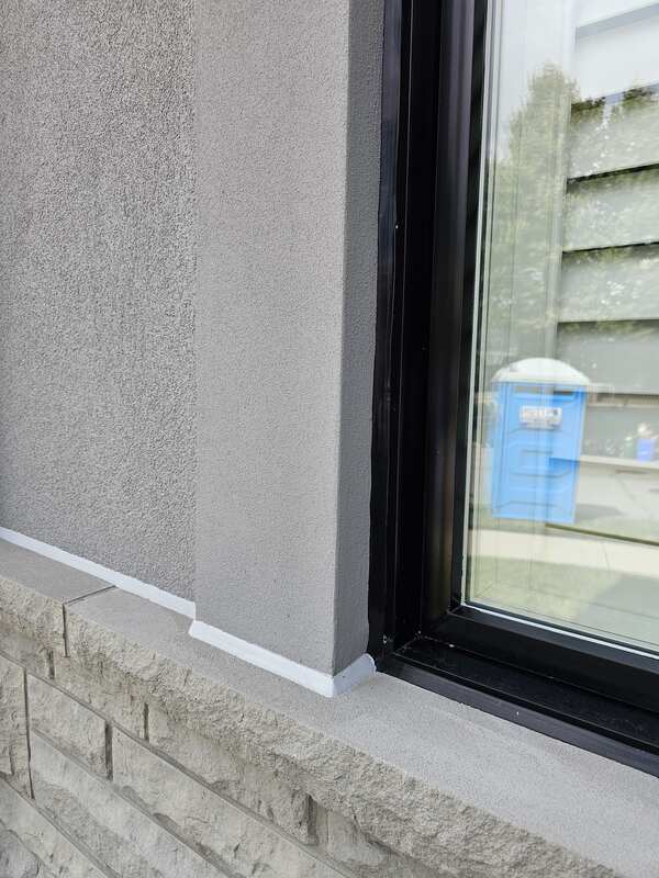 Close-up of a completed black silicone window seal meeting a freshly tooled white silicone expansion joint seal where stucco cladding meets a stone masonry ledger.