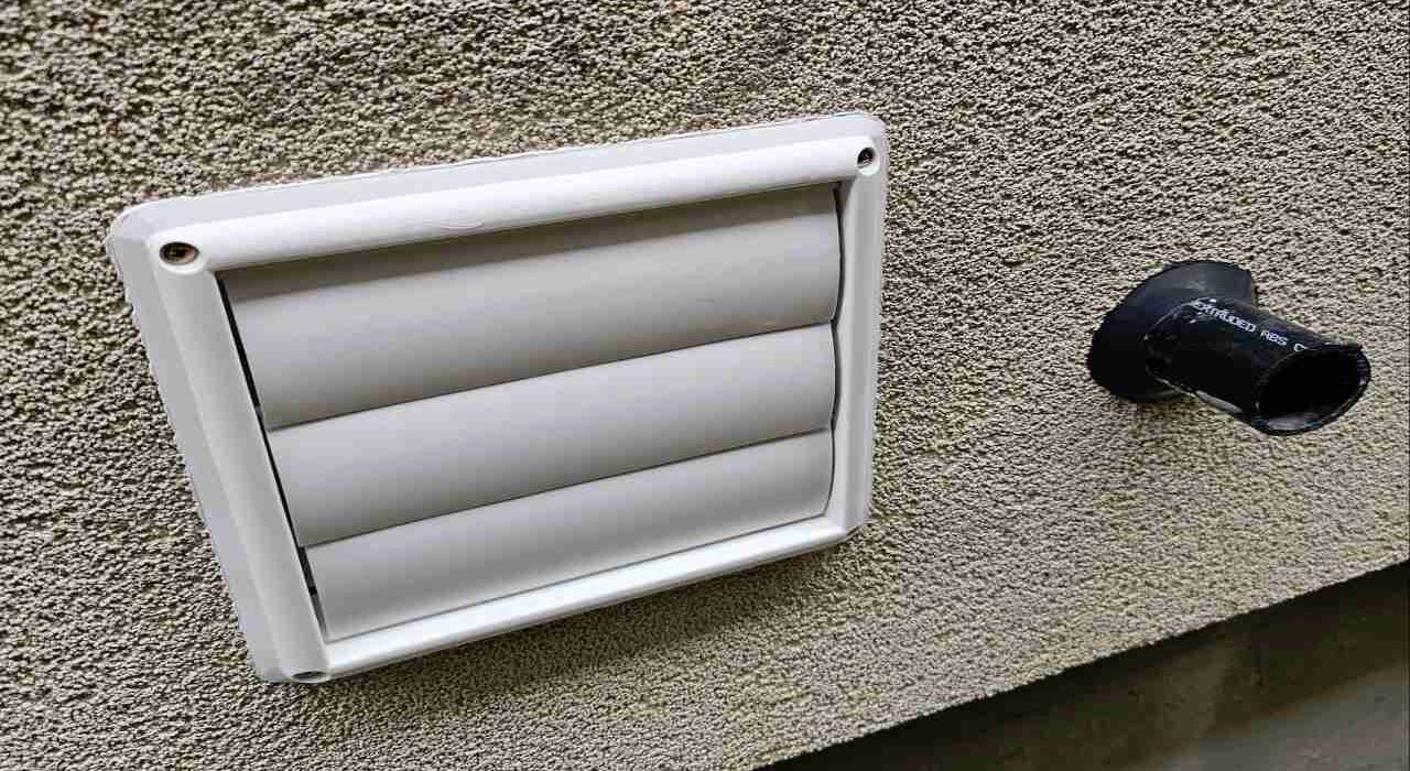 Side angle view of a white louvered dryer exhaust vent and black utility pipe mounted on a textured tan stucco exterior wall.