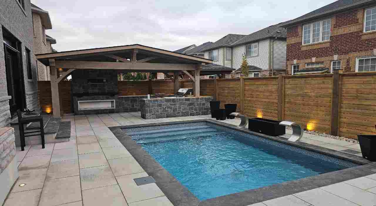 Back patio view of a luxury inground swimming pool featuring stone coping joints, large patio slabs, a covered outdoor kitchen, and a wooden privacy fence.