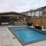 Back patio view of a luxury inground swimming pool featuring stone coping joints, large patio slabs, a covered outdoor kitchen, and a wooden privacy fence.