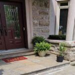 Front entry porch of a luxury house featuring a dark wood double door, natural stone masonry walls, flagstone landing slabs, and large structural columns.