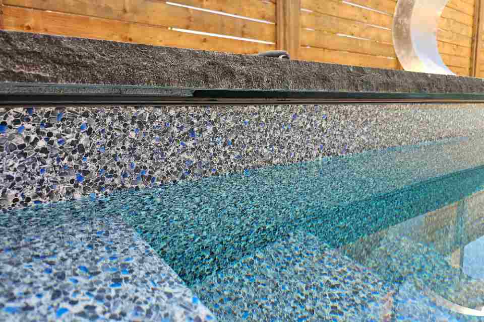 Extreme close-up of a perfectly tooled black silicone expansion joint seal directly beneath a dark stone coping edge and above a speckled blue pool tile waterline.