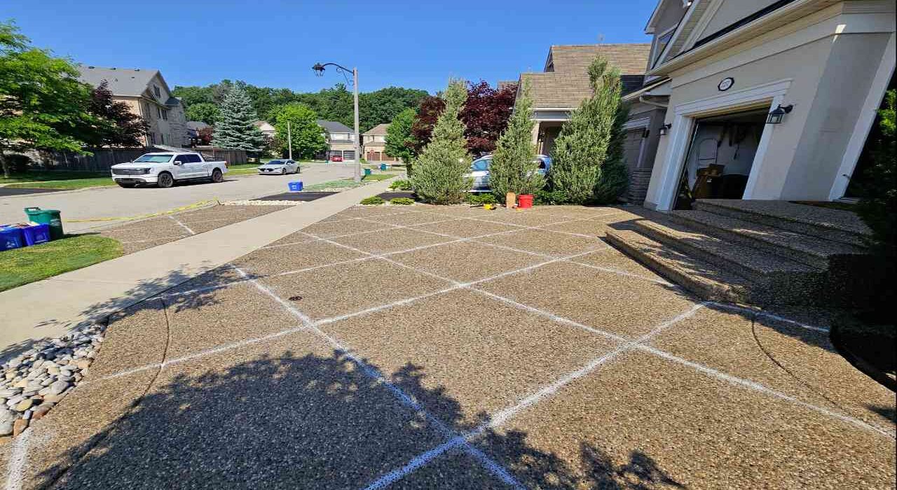 Wide exterior view of a luxury suburban house featuring a large exposed aggregate concrete driveway with a diamond-patterned grid layout of prepared control joints under a bright blue sky.