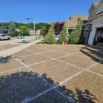Wide exterior view of a luxury suburban house featuring a large exposed aggregate concrete driveway with a diamond-patterned grid layout of prepared control joints under a bright blue sky.