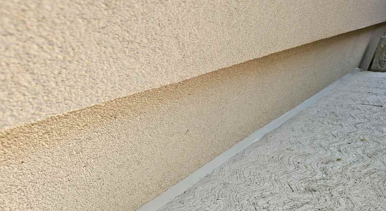 Long angled view of a flawlessly tooled tan silicone expansion joint seal along a modern residential stucco foundation wall and textured concrete walkway.