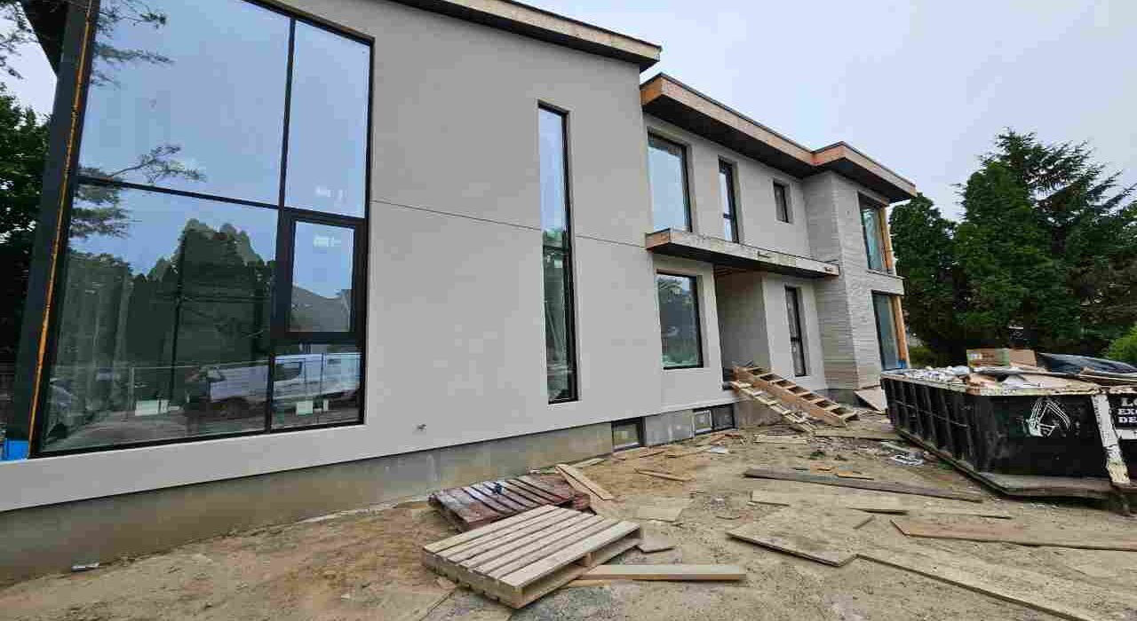 Front exterior view of a modern multi-story stucco home under construction featuring large black window frames undergoing professional sealing services.
