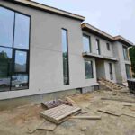 Front exterior view of a modern multi-story stucco home under construction featuring large black window frames undergoing professional sealing services.