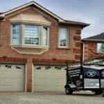Front view of a two-story brick home with a branded Proper Caulking service trailer parked in the driveway during a residential window sealing project.