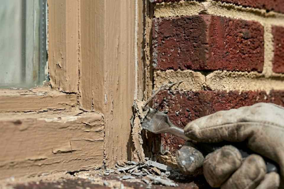 Proper Caulking technician carefully removing old brittle caulking out of a window joint away from the wood trim on a brick home in Burlington.