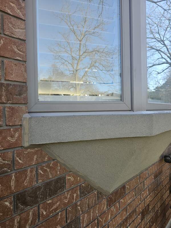 A close-up view of a brick house showing completed bay window exterior caulking transitioning between a precast concrete sill, a vinyl frame, and a stucco base.