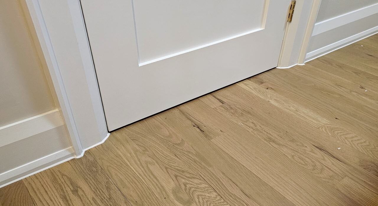 A low floor-level view of a brick house showing a completed baseboard and door casing caulking job against hardwood flooring.