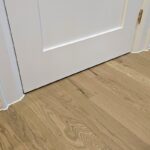 A low floor-level view of a brick house showing a completed baseboard and door casing caulking job against hardwood flooring.