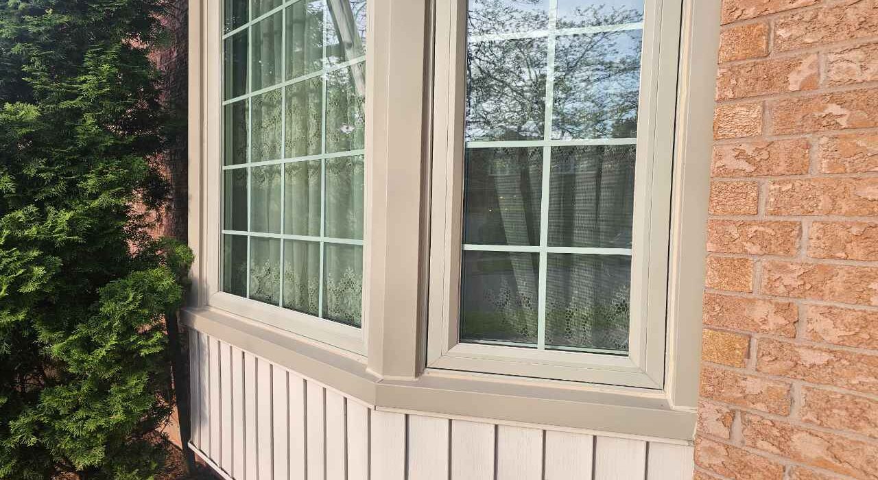 A wide close-up landscape view of a completed bay window exterior caulking job showing a flawless white vertical sealant bead on a 45-degree corner mullion joint.