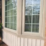 A wide close-up landscape view of a completed bay window exterior caulking job showing a flawless white vertical sealant bead on a 45-degree corner mullion joint.