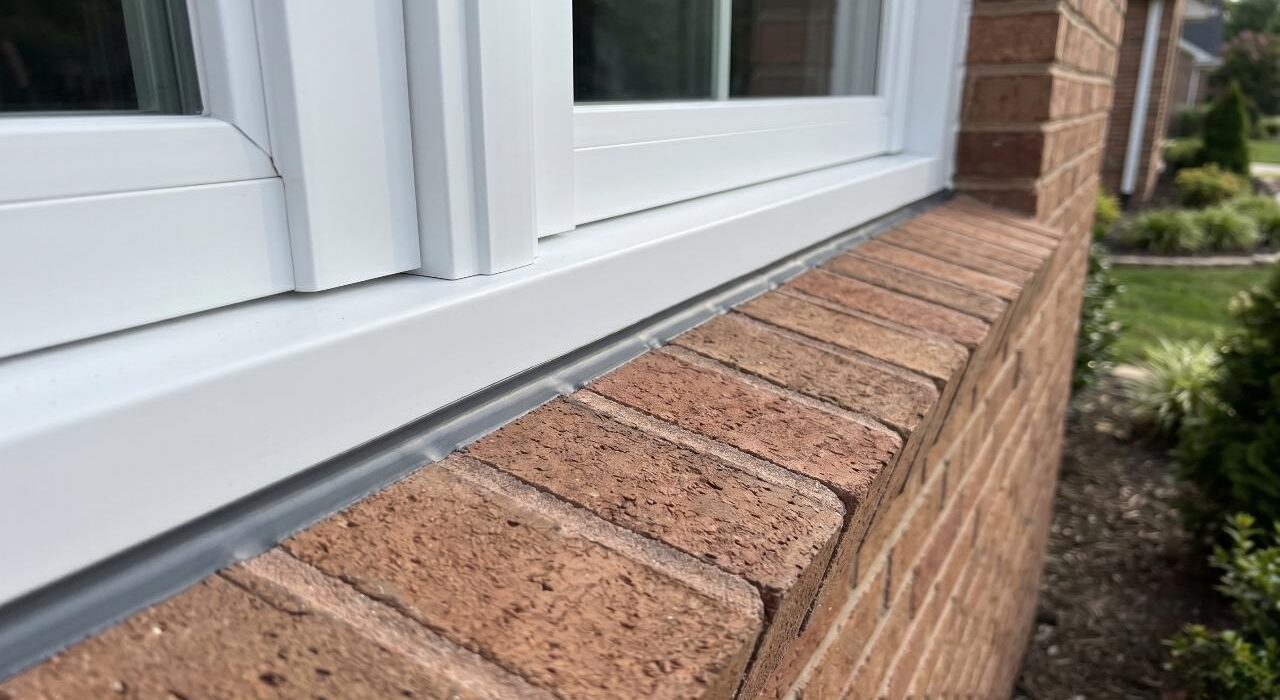 A wide close-up landscape view of a completed brick window sill caulking job showing a flawless horizontal sealant bead on an angled rowlock masonry ledge beneath a white vinyl frame.