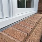 A wide close-up landscape view of a completed brick window sill caulking job showing a flawless horizontal sealant bead on an angled rowlock masonry ledge beneath a white vinyl frame.