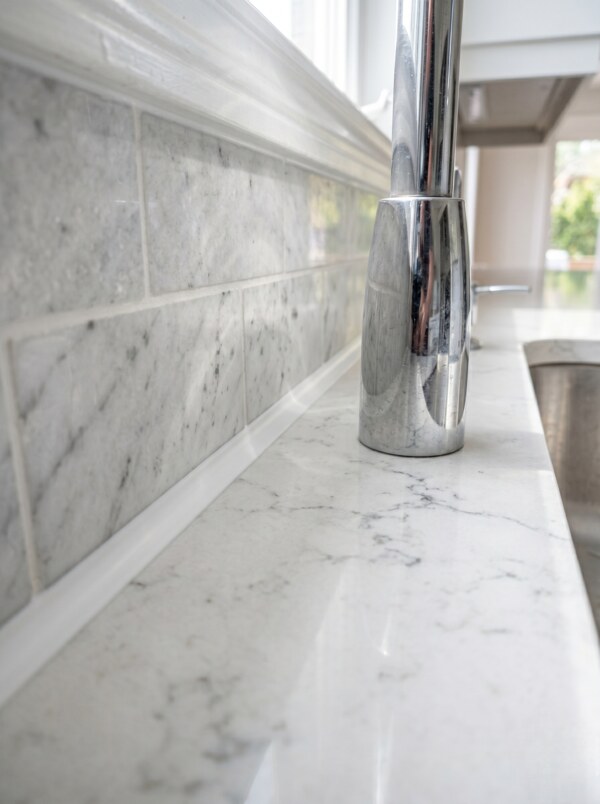 A vertical close-up view of a kitchen counter showing a completed backsplash sealing job with a flawless thin white silicone bead along a quartz countertop joint.