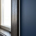 A close-up landscape view of a completed interior window sealing job showing a crisp white sealant bead contrasting against dark oak casing and a navy wall.