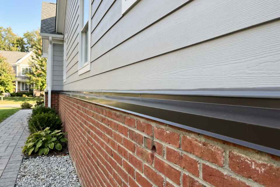 A wide 4:3 field photograph looking down a completed horizontal water table transition showing a flawless grey sealant bead bonding bronze metal drip cap flashing to grey siding and a red brick foundation wall.