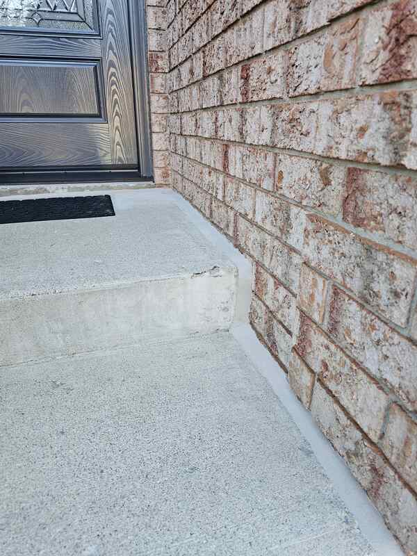 Looking at a completed porch-to-house expansion joint showing a crisp grey sealant bead bonding a concrete entryway staircase step to a red clay brick wall.