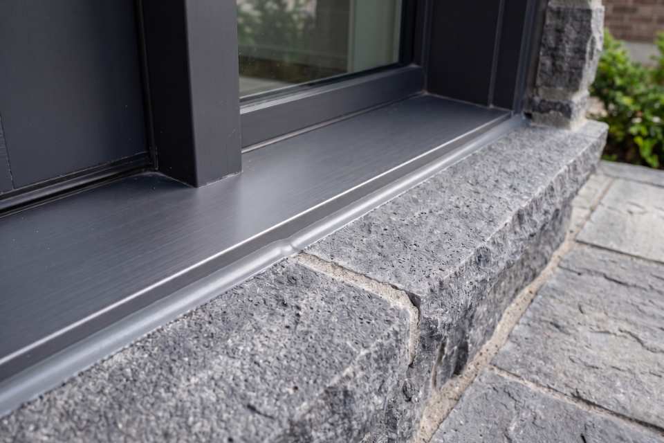 Looking down at a completed exterior door threshold caulking job showing a flawless gray silicone bead sealing a horizontal charcoal metal sill to a clean stone porch landing block.