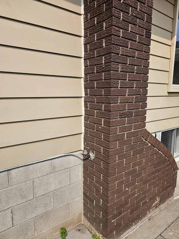 A wide view of a completed exterior caulking job showing a white polymer bead sealing tan lap siding boards and a brick chimney to a grey concrete block foundation wall.