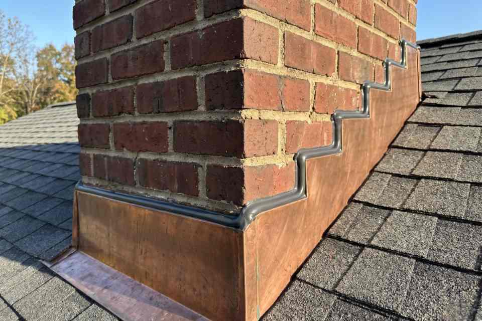 A close-up view of a completed chimney counter-flashing caulking job showing a flawless grey polyurethane bead sealing a stepped copper flashing track to a red brick masonry chimney wall.