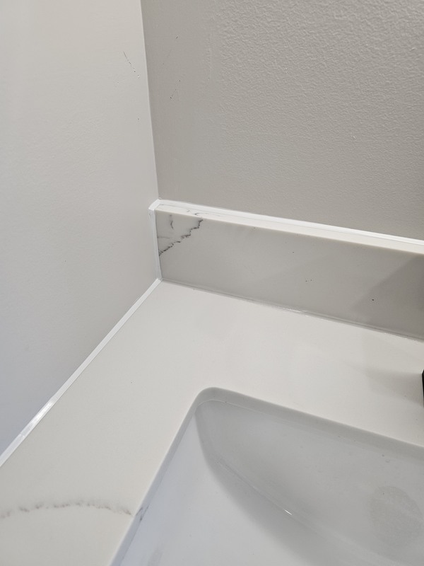 A close-up view of a washroom vanity showing crisp white silicone caulking joints sealing a quartz countertop to a matching stone backsplash and painted walls.