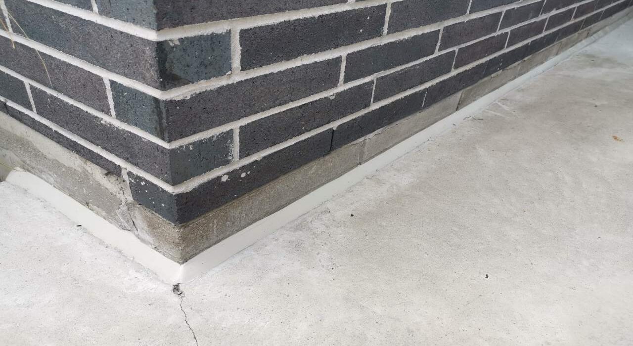 A sharp, sloped white polyurethane caulking bead sealing a horizontal concrete sidewalk pad to a dark grey brick masonry foundation wall.