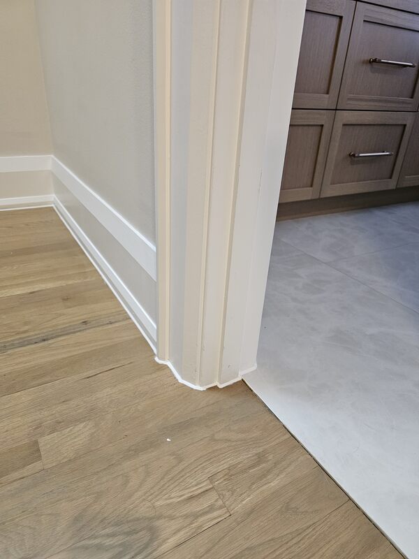 A close-up view of an interior doorway showing fresh white caulking wrapping a door casing profile and baseboard trim between hardwood and tile floors.