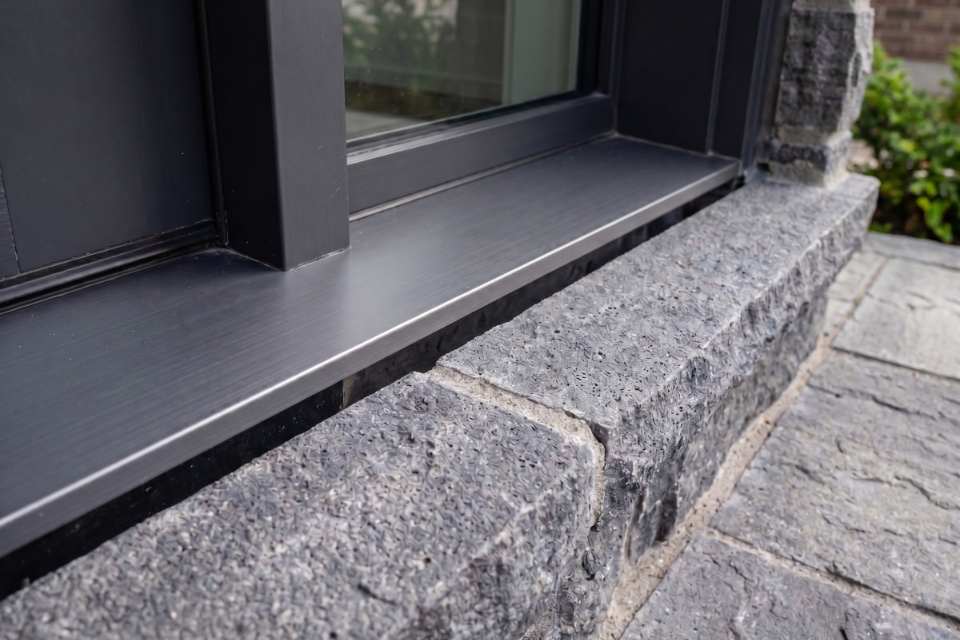 Looking at an empty horizontal joint channel between a black aluminum door frame and rough grey split-face stone masonry after old caulking removal.