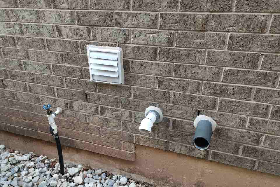 A close-up view of an exterior brick wall showing four utility penetrations including a louvered exhaust vent, a water hose spigot, and two PVC pipes requiring exterior utility caulking.