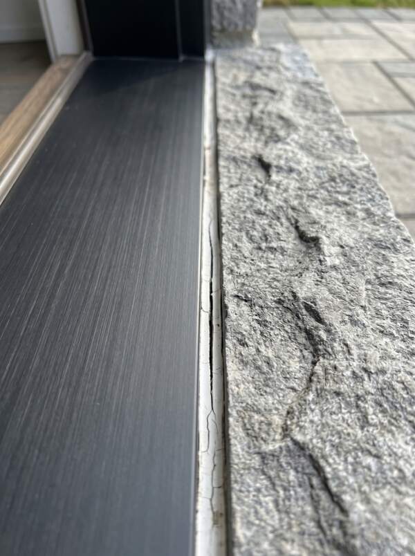 Failed exterior door caulking showing a thin, narrow white retail sealant splitting away from a black aluminum door sill and a clean grey stone porch landing.