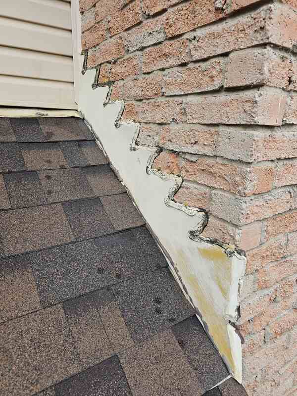 Failed exterior caulking showing a dirty, cracked, and dried-out white sealant line on a stepped metal roof flashing track against a red brick wall.