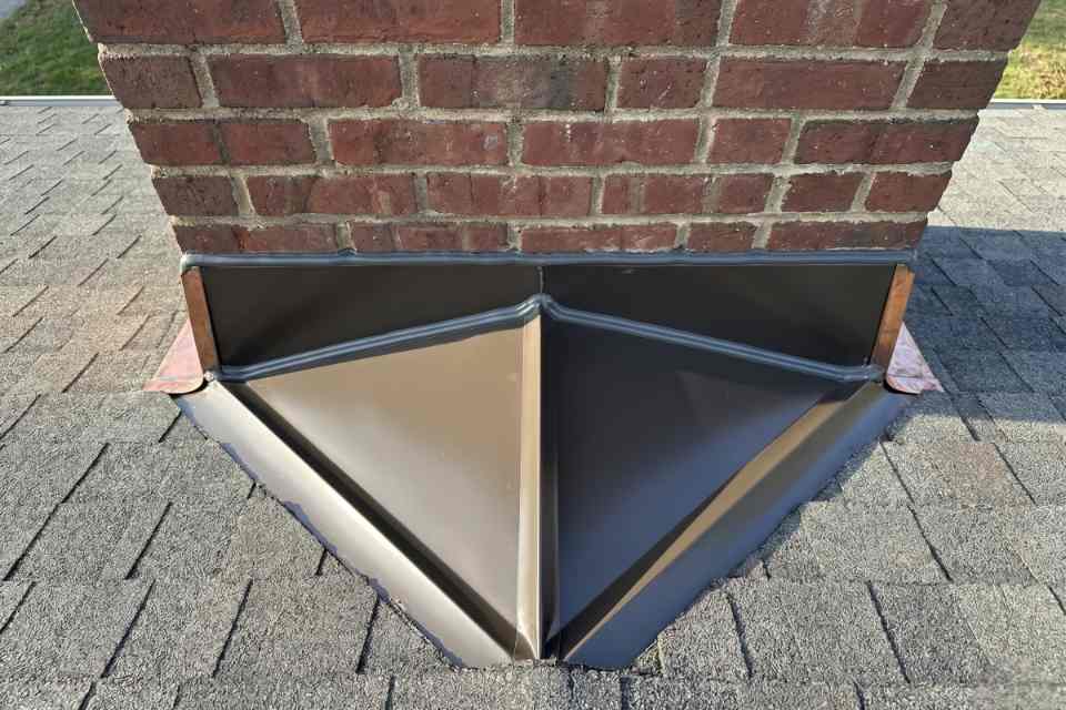 An overhead view looking down at a completed chimney cricket flashing seal showing a flawless grey sealant bead bonding a dark bronze metal saddle to a red brick chimney wall.