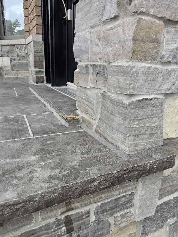 Completed exterior caulking showing a crisp grey commercial sealant bead bonding a flagstone porch pad and door threshold sill to a rugged stone pillar wall.
