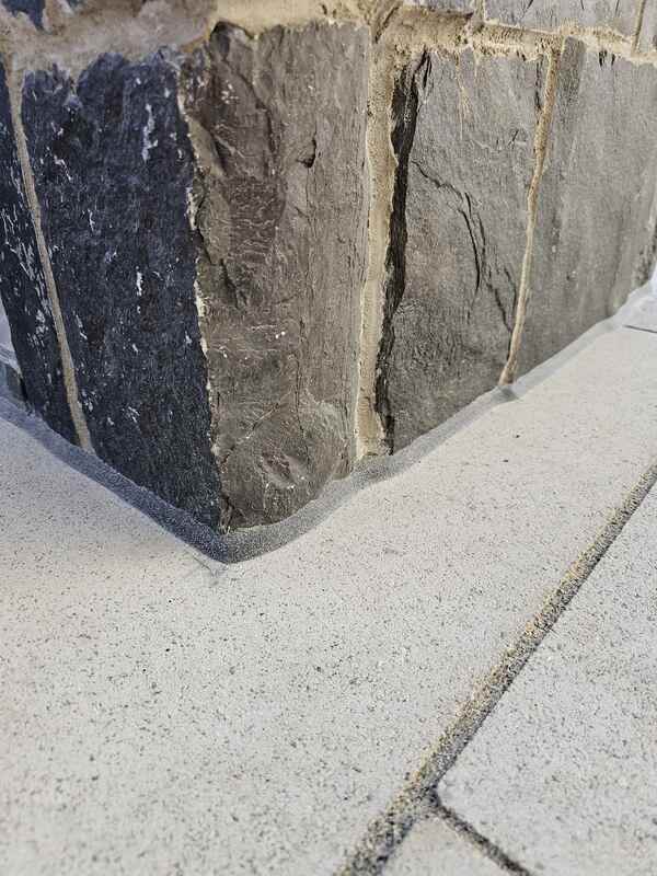 Looking closely at a completed porch expansion joint showing a clean grey commercial sealant bead bonding a concrete slab to a textured dark stone masonry wall.